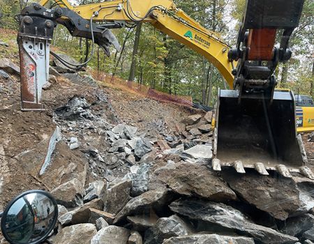 An excavator with a jackhammer attachment breaking rocks on a hillside with an orange construction barrier.