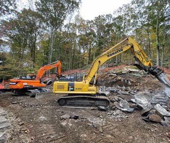 Two excavators, one orange, one yellow, are breaking rocks on a construction site. Trees in the background.