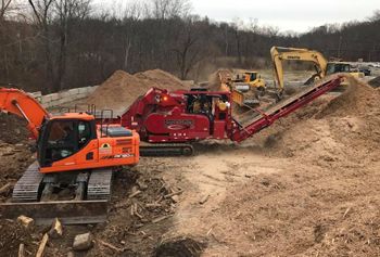 Construction site with an orange excavator and a red grinder processing wood chips; a yellow excavator in the background.