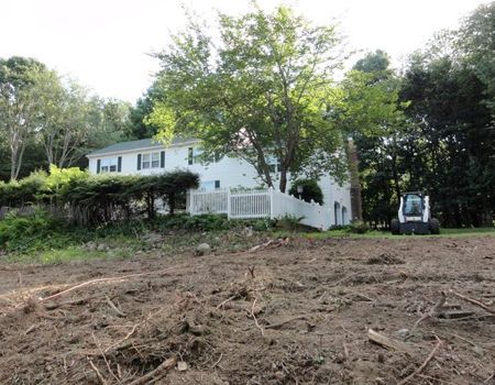Cleared land in front of a white house with a Bobcat. Brush and debris cover the foreground.