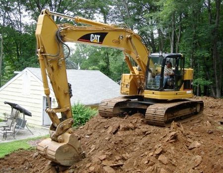 Yellow Caterpillar excavator digging into a pile of dirt near a house. A person is visible in the cab operating the machine.
