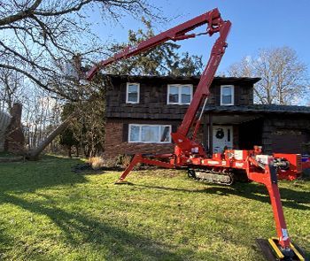 A red tree-trimming lift is positioned in a grassy yard near a two-story brick house, with the lift's arm