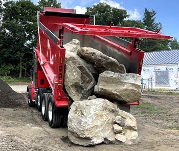 Red dump truck unloading large boulders on a dirt construction site, trees and a building in the background.