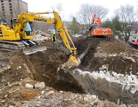 Two excavators dig into a pile of debris and earth, one yellow, one orange, at a construction site.