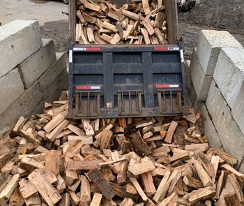 A truck bed full of split firewood is being dumped between two concrete barriers.