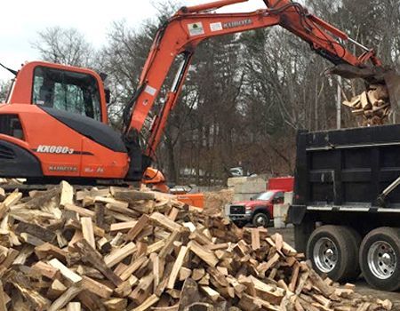 An orange excavator loads firewood into a black dump truck. A large pile of wood sits on the ground in a daylight setting.
