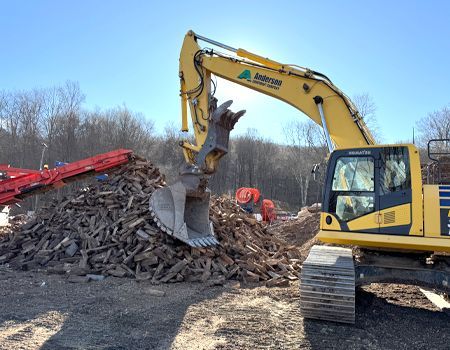 A yellow excavator with a claw-like attachment scoops up wood debris, with a red conveyor belt and trees in the background.