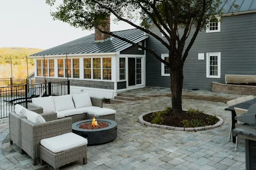 Patio with gray wicker seating, fire pit, and tree beside a gray house with sunroom and stone pavers