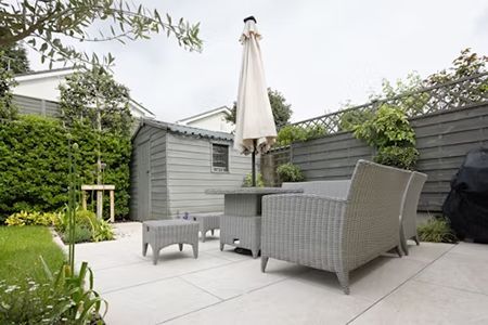 A gray patio with woven furniture, a closed umbrella, and a shed, surrounded by greenery and a fence.