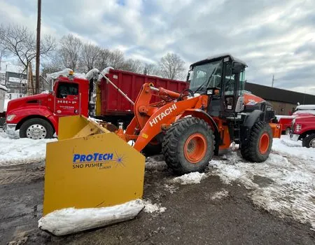 A hitachi snow plow is parked in the snow next to a red truck.