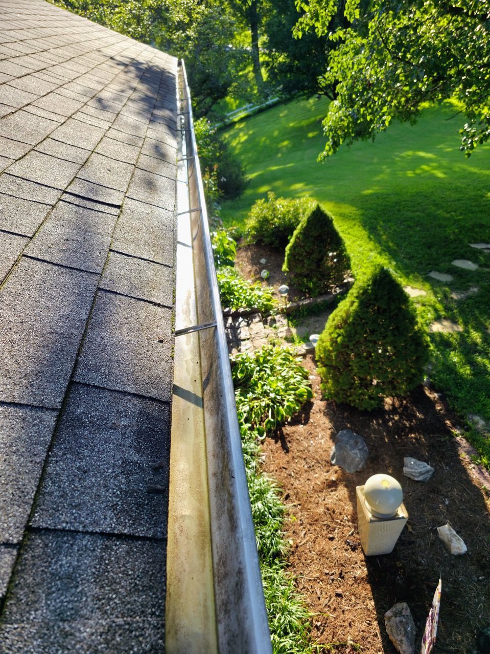 Roof gutter beside a lush green garden bed with shrubs and stones in sunlight