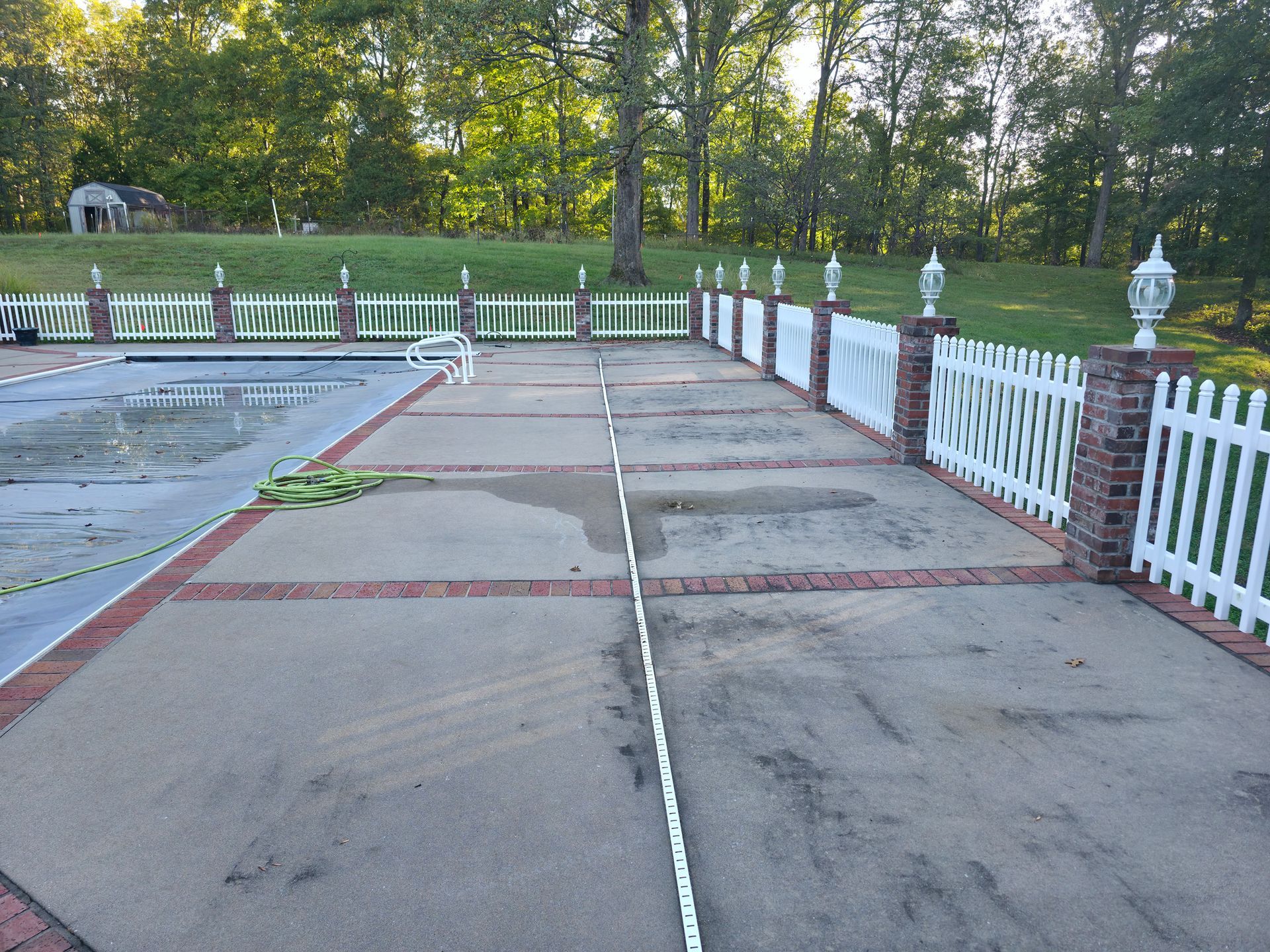 Empty outdoor walkway beside a pool, with white fences and green trees in the background.