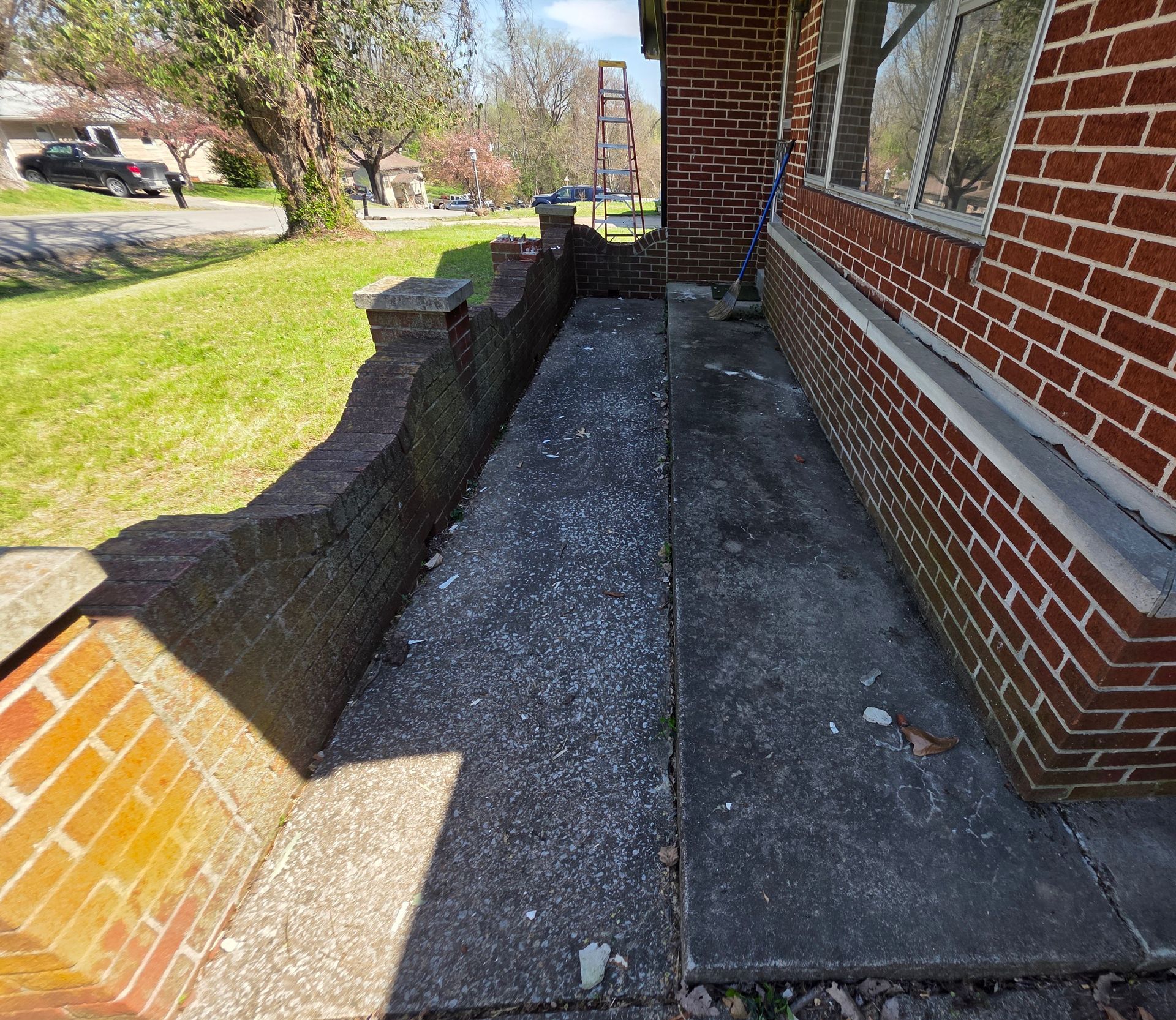 Narrow concrete side yard beside a brick house, with low brick wall and grass lawn in sunlight