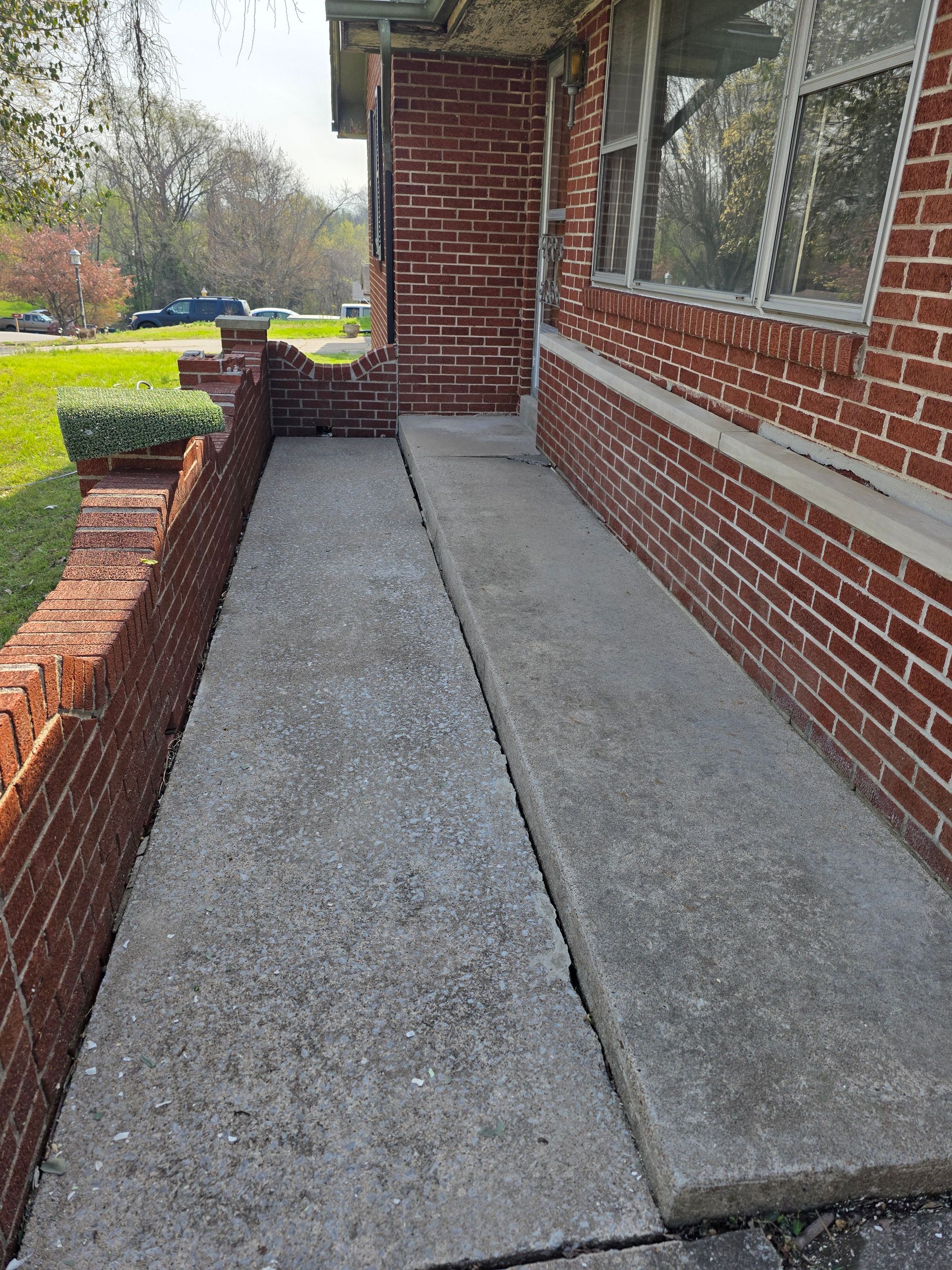 Concrete walkway beside a red brick house, bordered by low brick planters and green lawn.