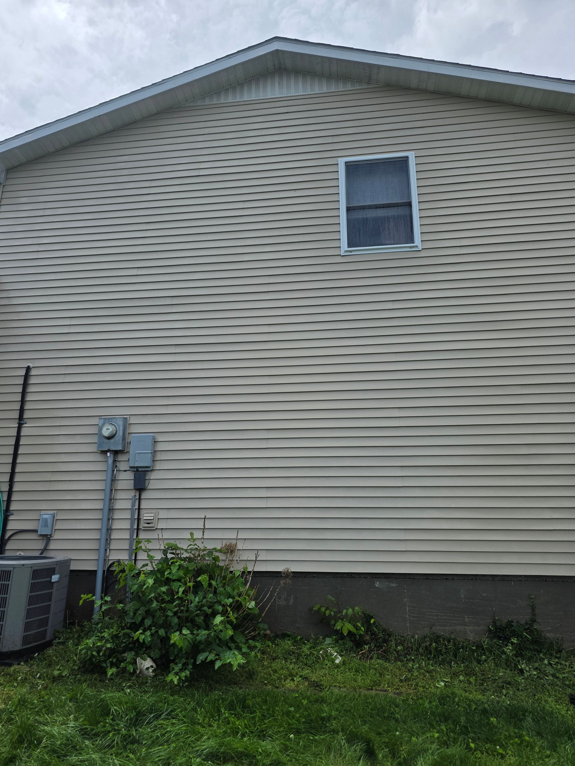 Side of a beige house with vinyl siding, a small window, utility meters, and grass in the foreground