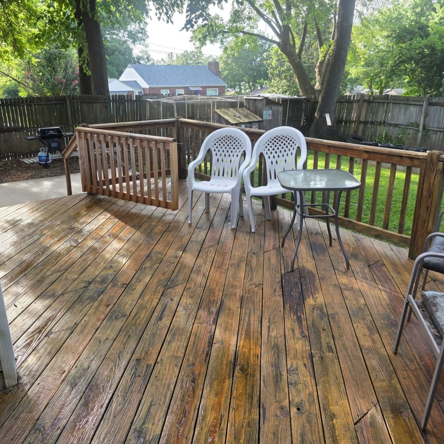 Wooden deck with rain puddles, two white patio chairs, and a small round table in a fenced backyard