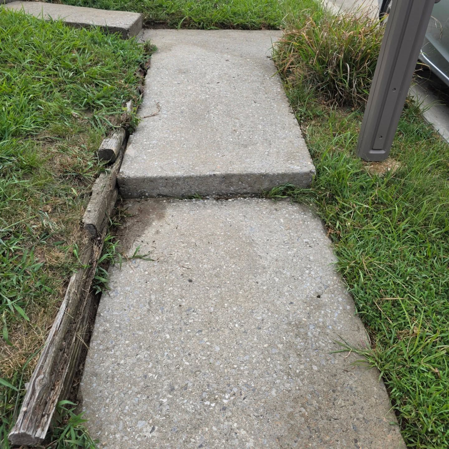 Cracked concrete sidewalk with a raised slab and a grassy edge beside a post