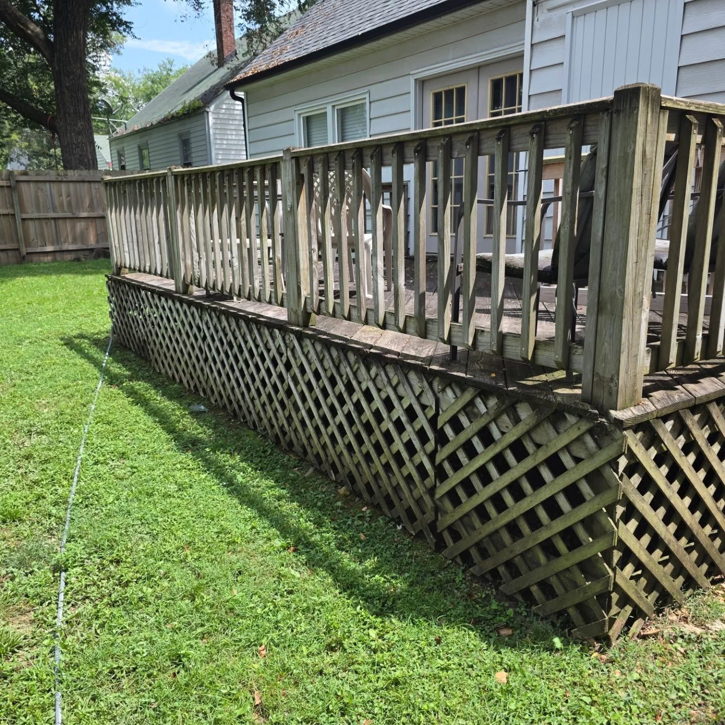Weathered wooden deck with leaning railing and lattice skirt beside a white house on a grassy yard.