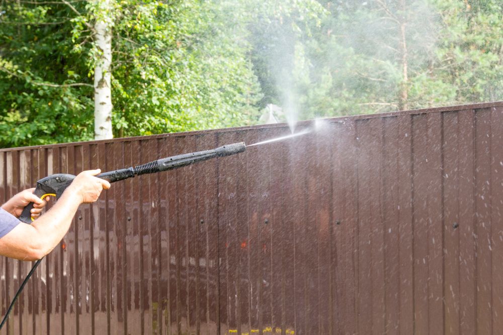 Person pressure-washes a brown wooden fence, spraying water over a backyard wall.