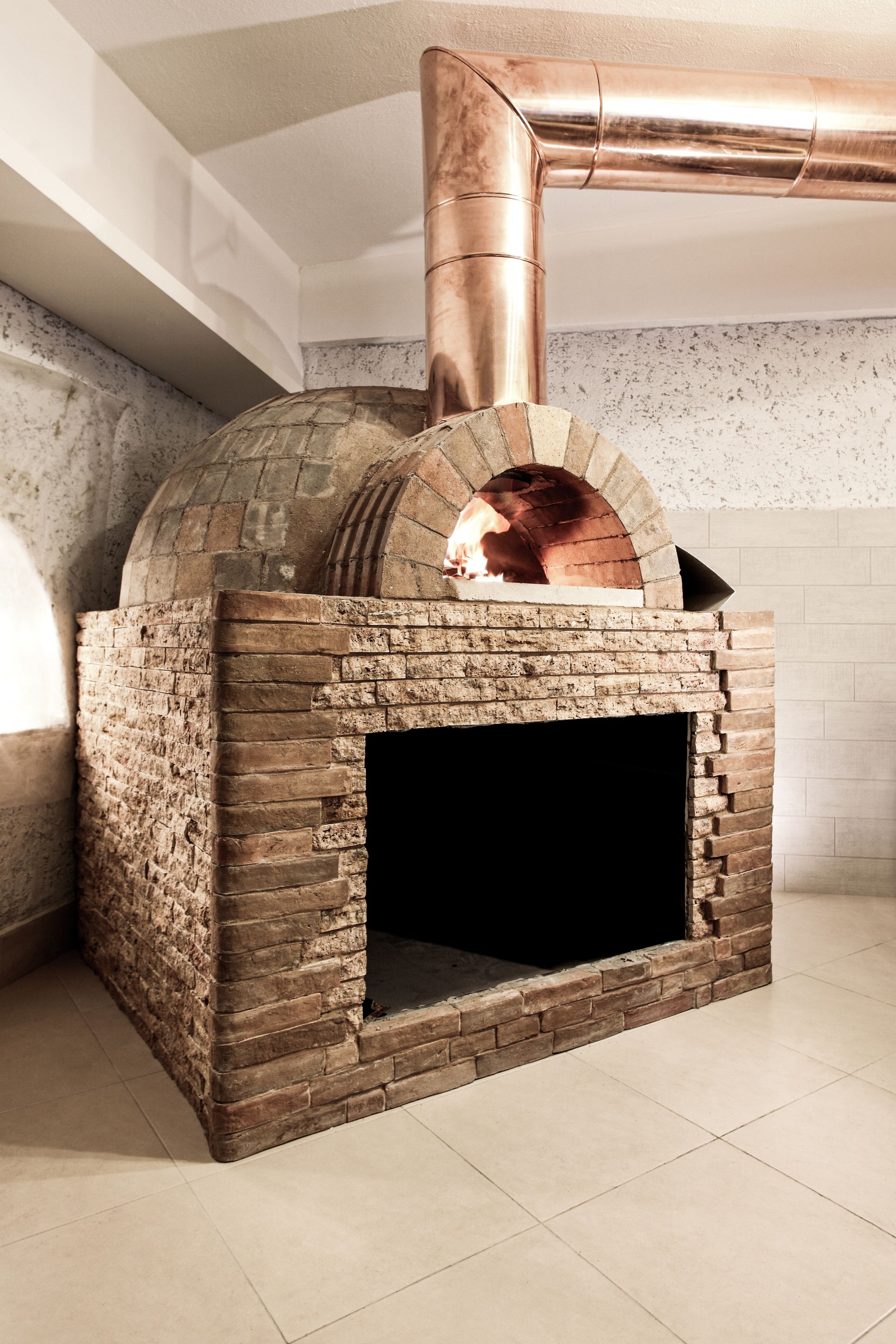 Wood-fired pizza oven with copper chimney, brick exterior, and open mouth, flames visible inside.