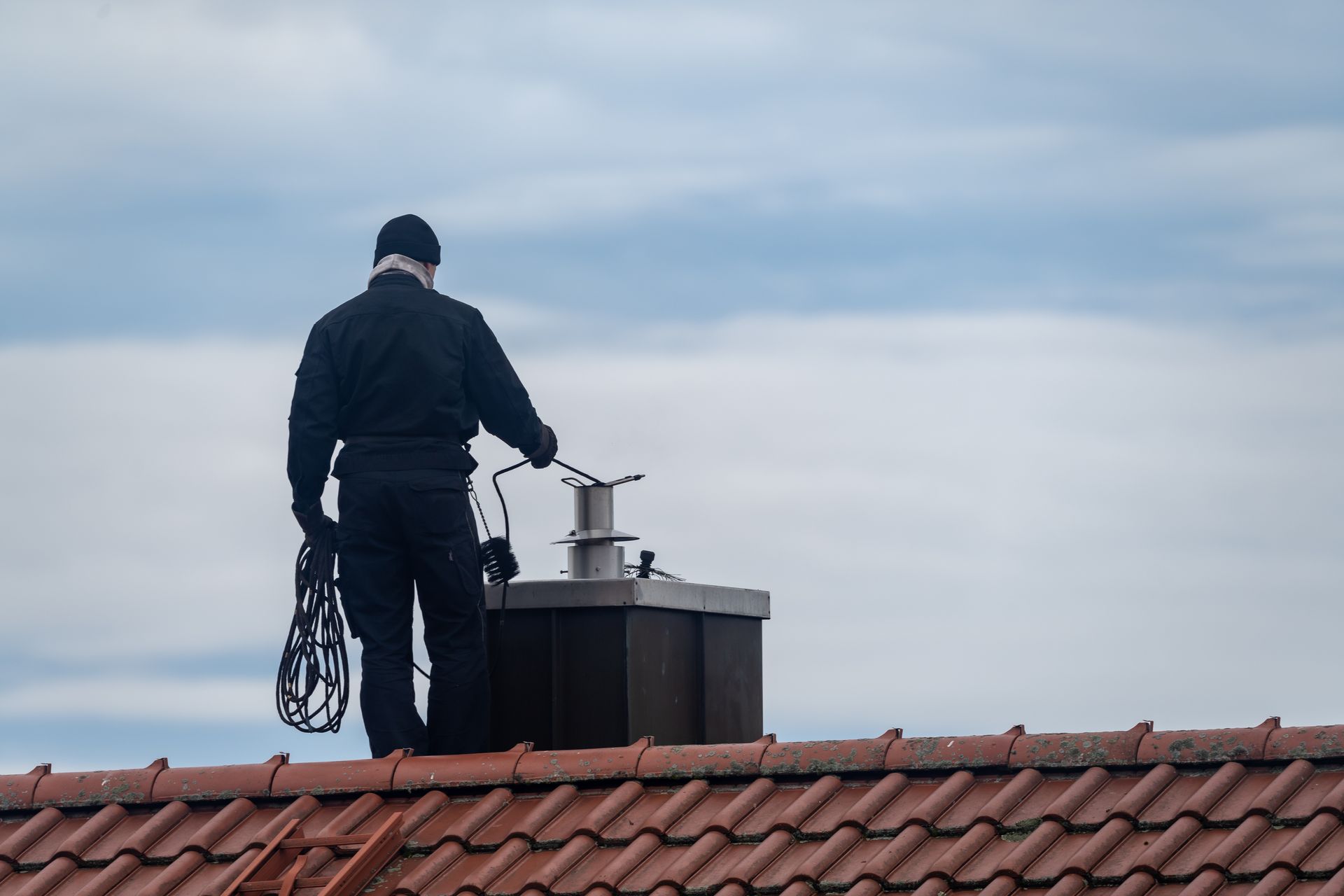 Chimney sweep on a rooftop, holding tools, working on chimney under a cloudy sky.