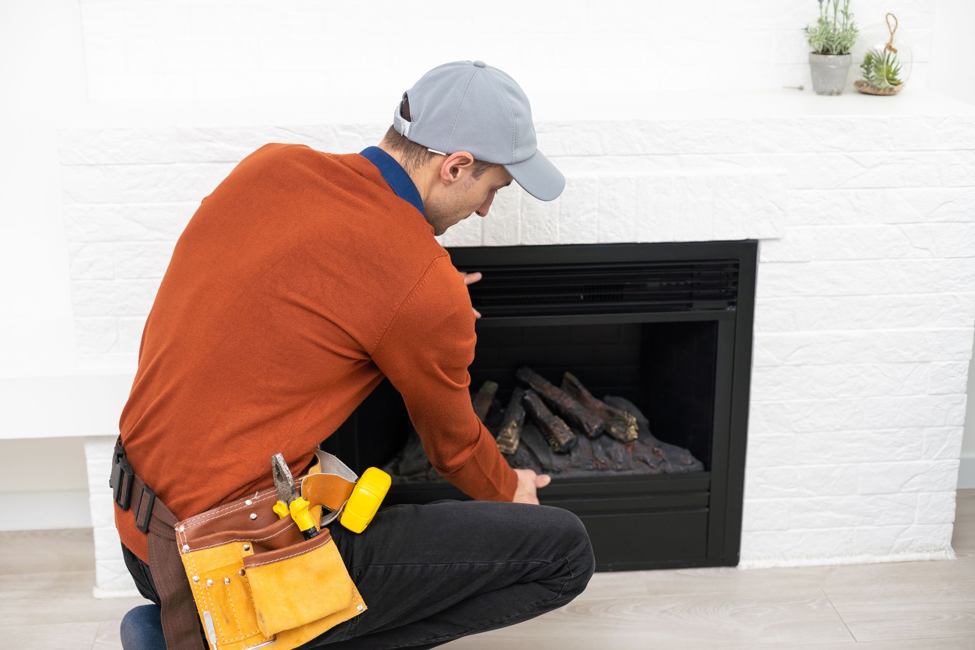 Man in orange shirt examines a fireplace, wearing a tool belt and cap.