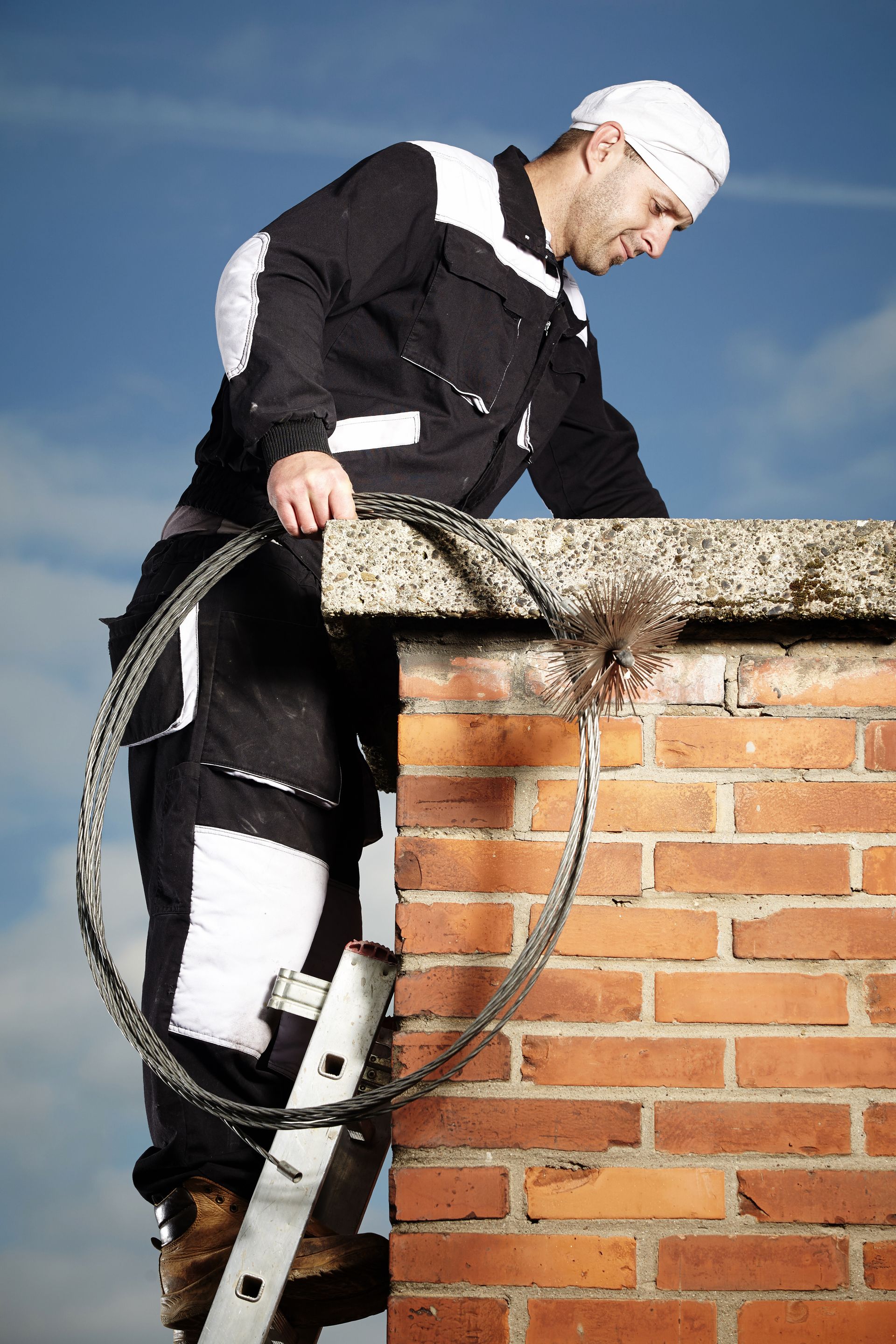 Man on ladder cleaning a brick chimney. Holding a coiled brush, blue sky.