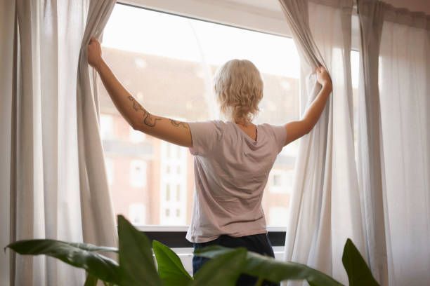Person holding open light-colored curtains in front of a large window with outdoor view.