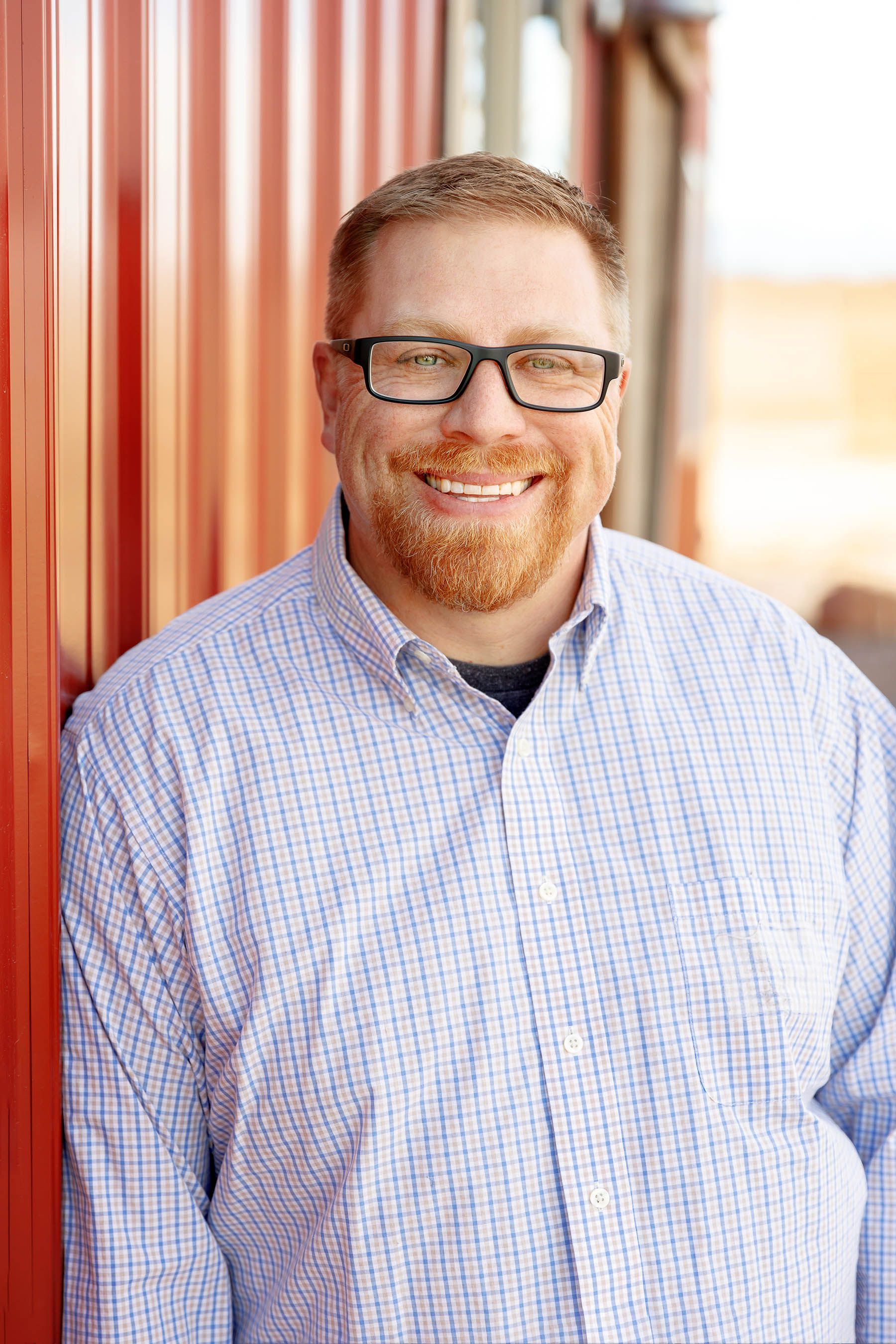 Smiling man in glasses and checkered shirt standing beside a red corrugated wall