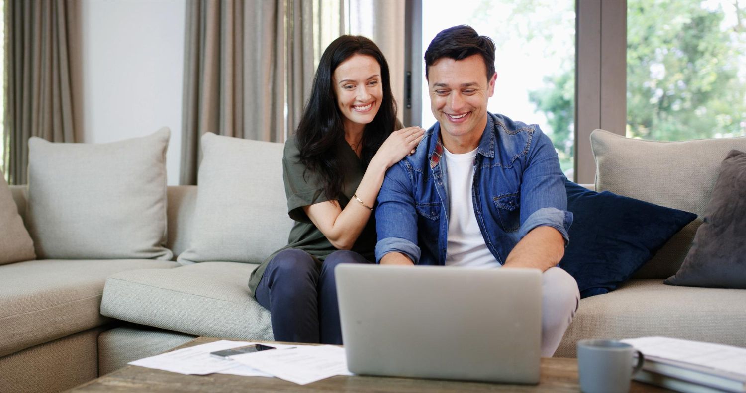 Two people smiling on a couch, looking at a laptop in a bright living room