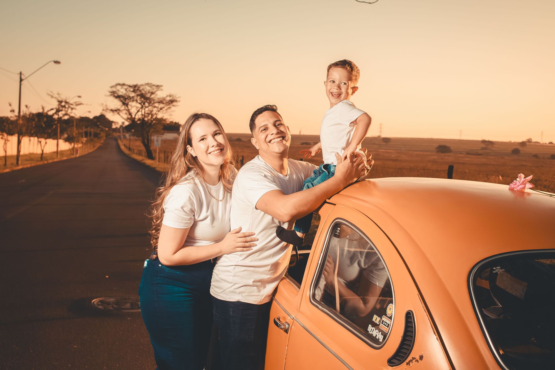 Three people smiling beside an orange car on a road at sunset