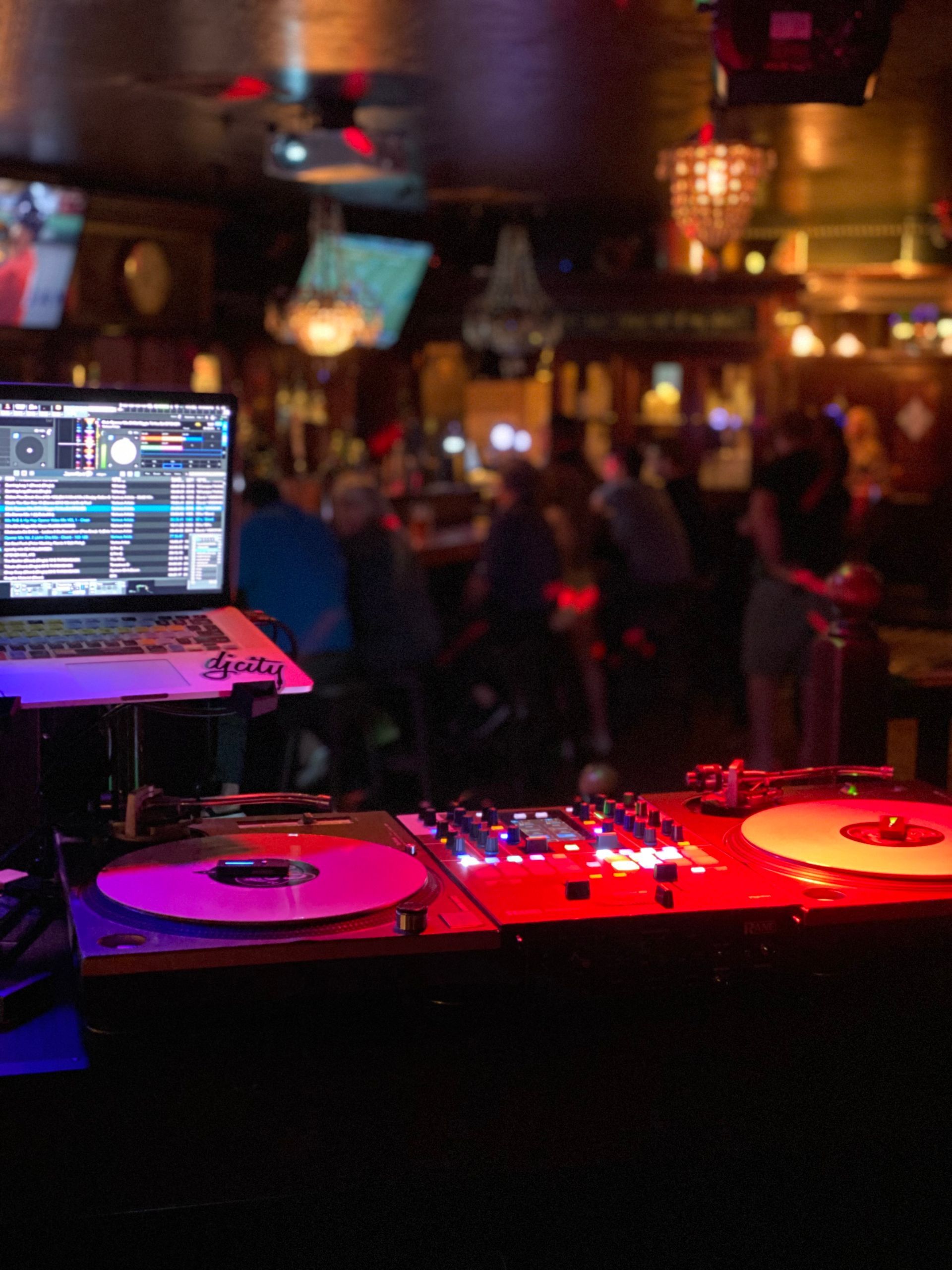 DJ setup with laptop, turntables, and mixer at a bar with patrons in the background. Red and purple lighting.