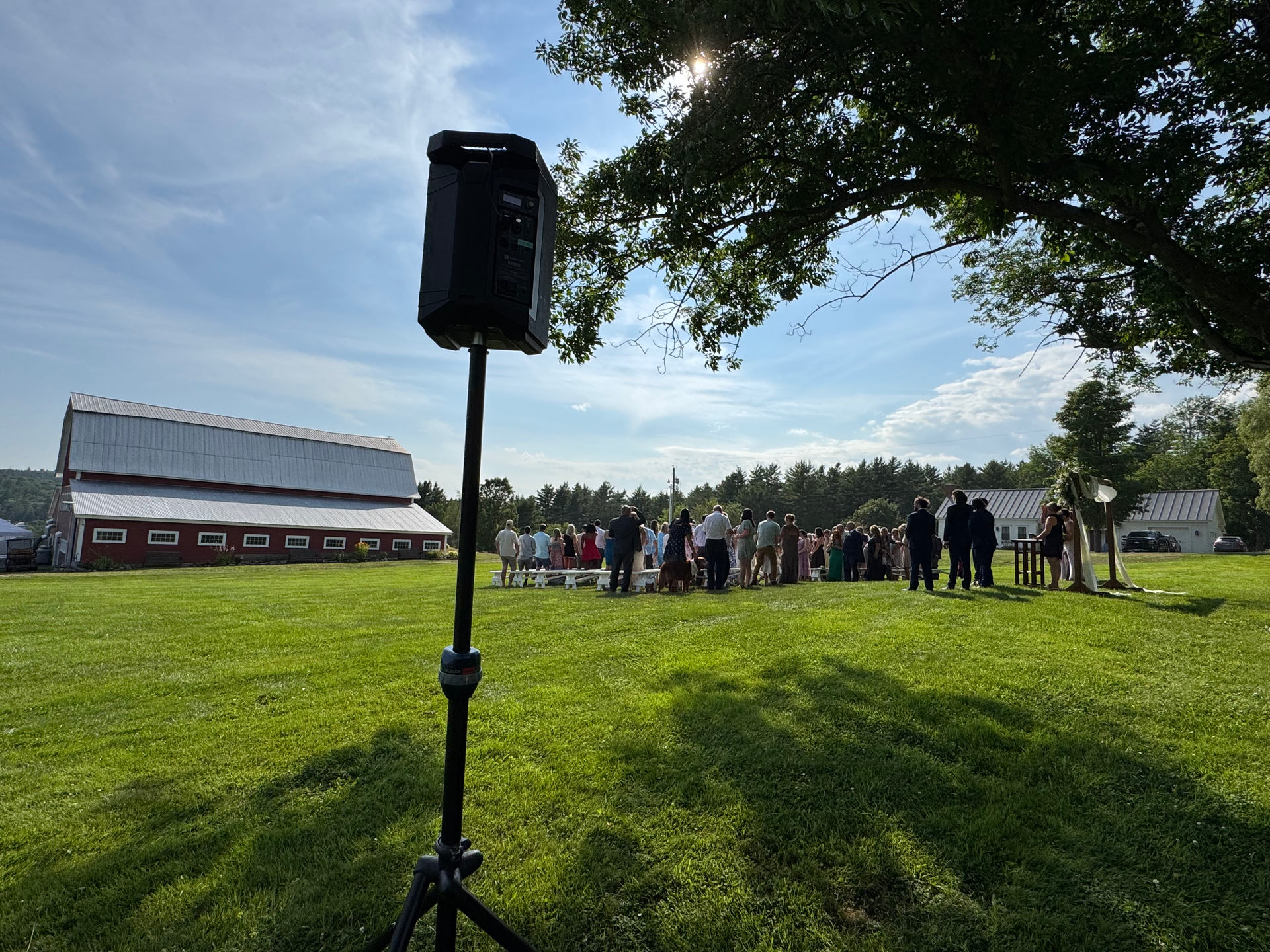 Speaker on tripod in grassy field, people gathered in the distance, red barn, sunny day.