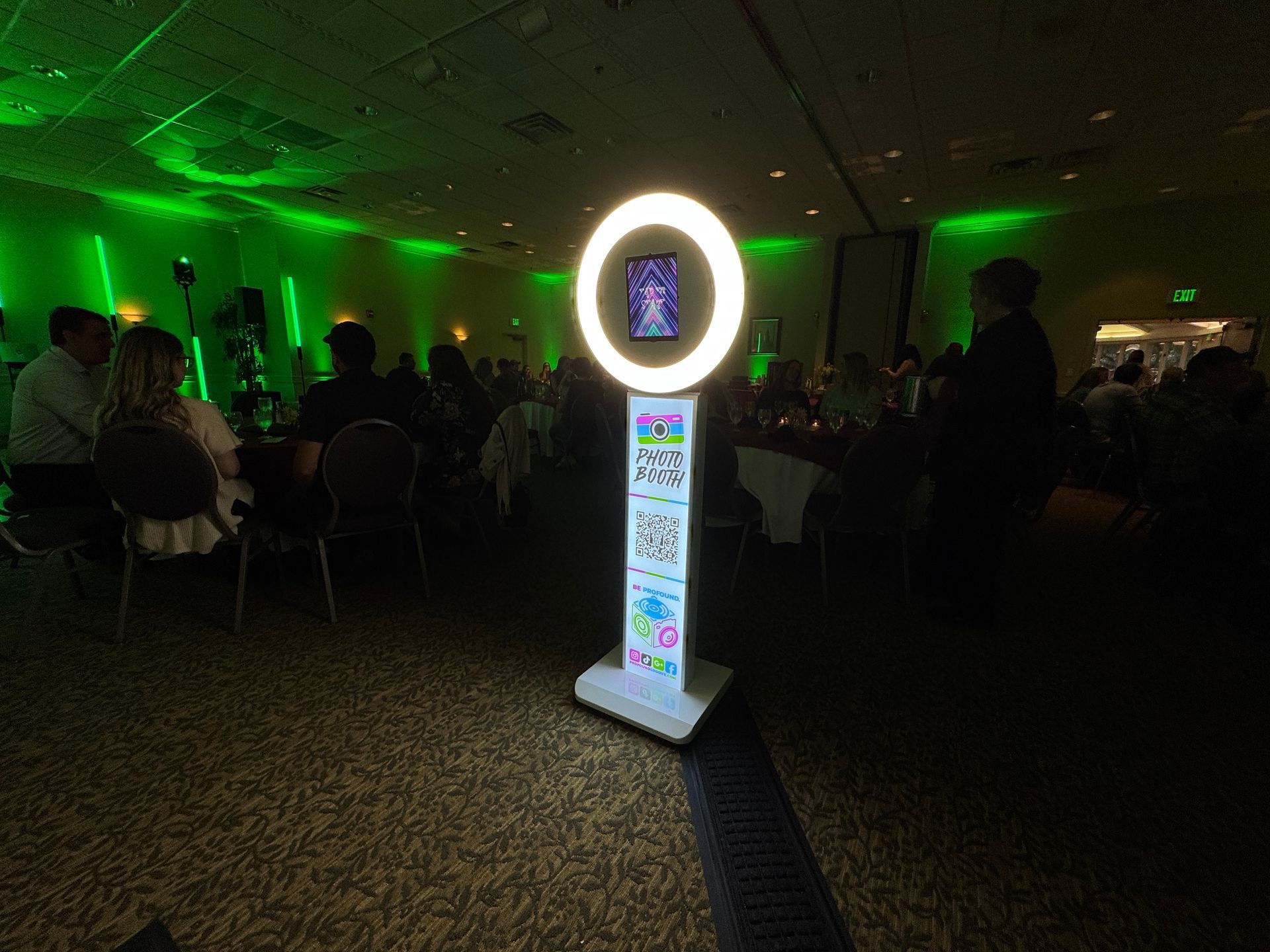 Photo booth with a ring light in a green-lit event hall; people sit at tables in the background.