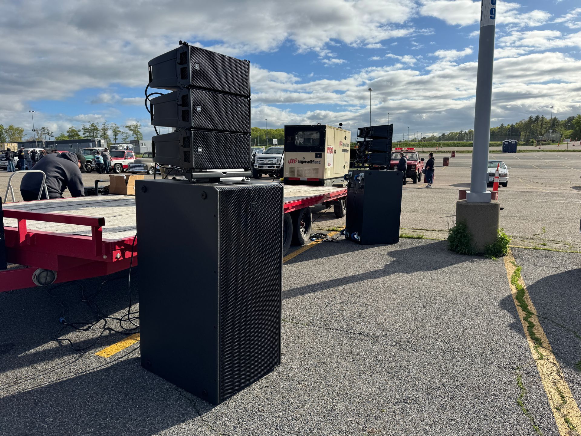 Black speakers stacked in a parking lot, on a red trailer, and on the ground, against a cloudy sky.