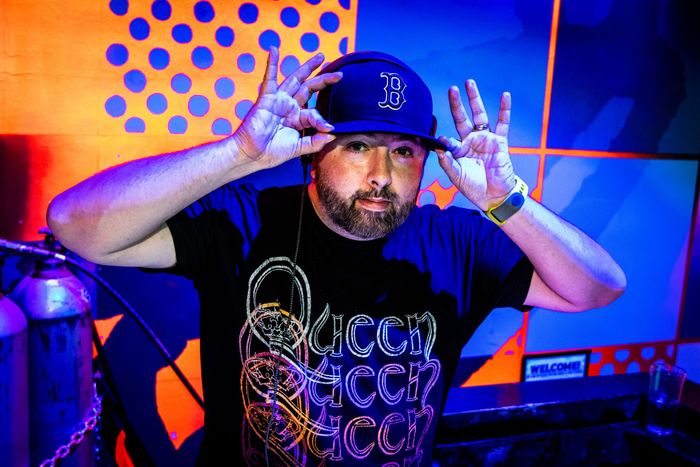 Man wearing a baseball cap and graphic t-shirt holds up his hands. Orange and blue background with polka dots.