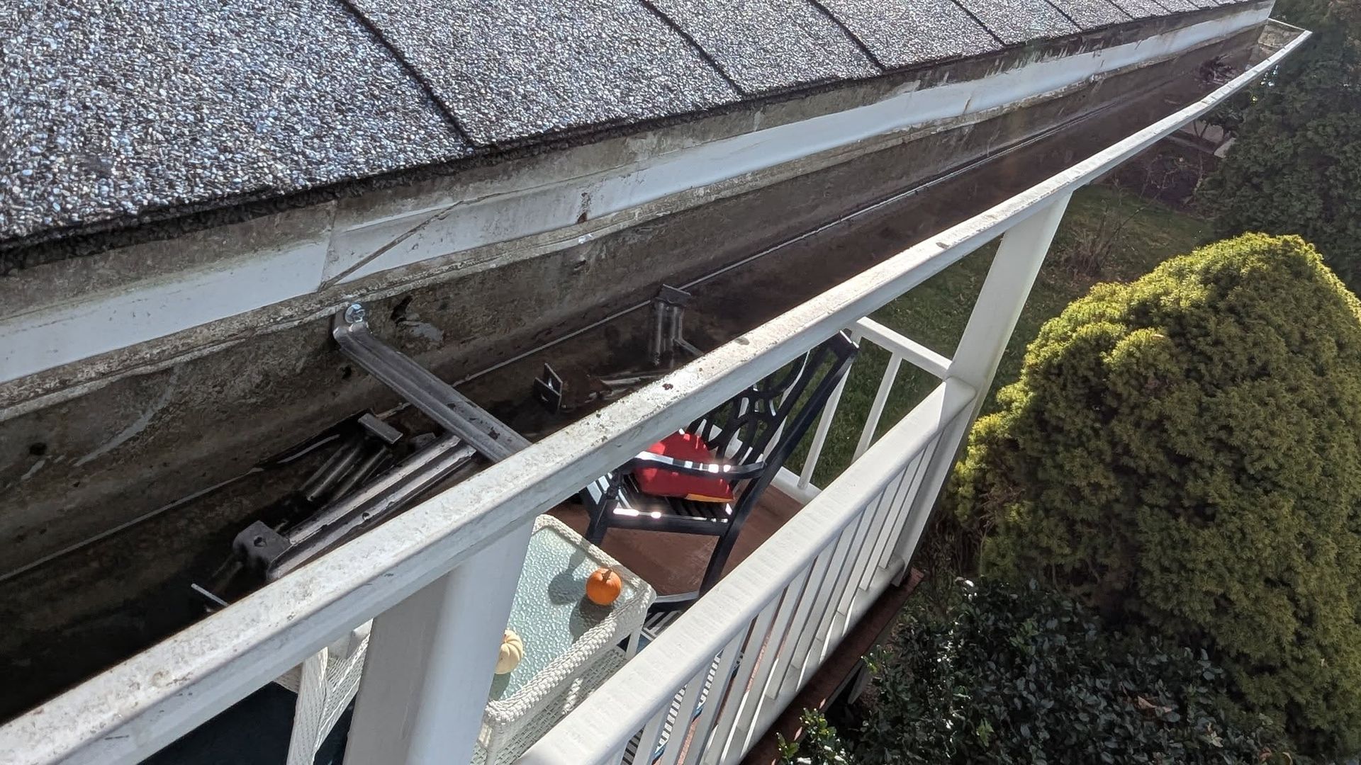 Roof gutter beside white railing, with a red car visible below and trees nearby.