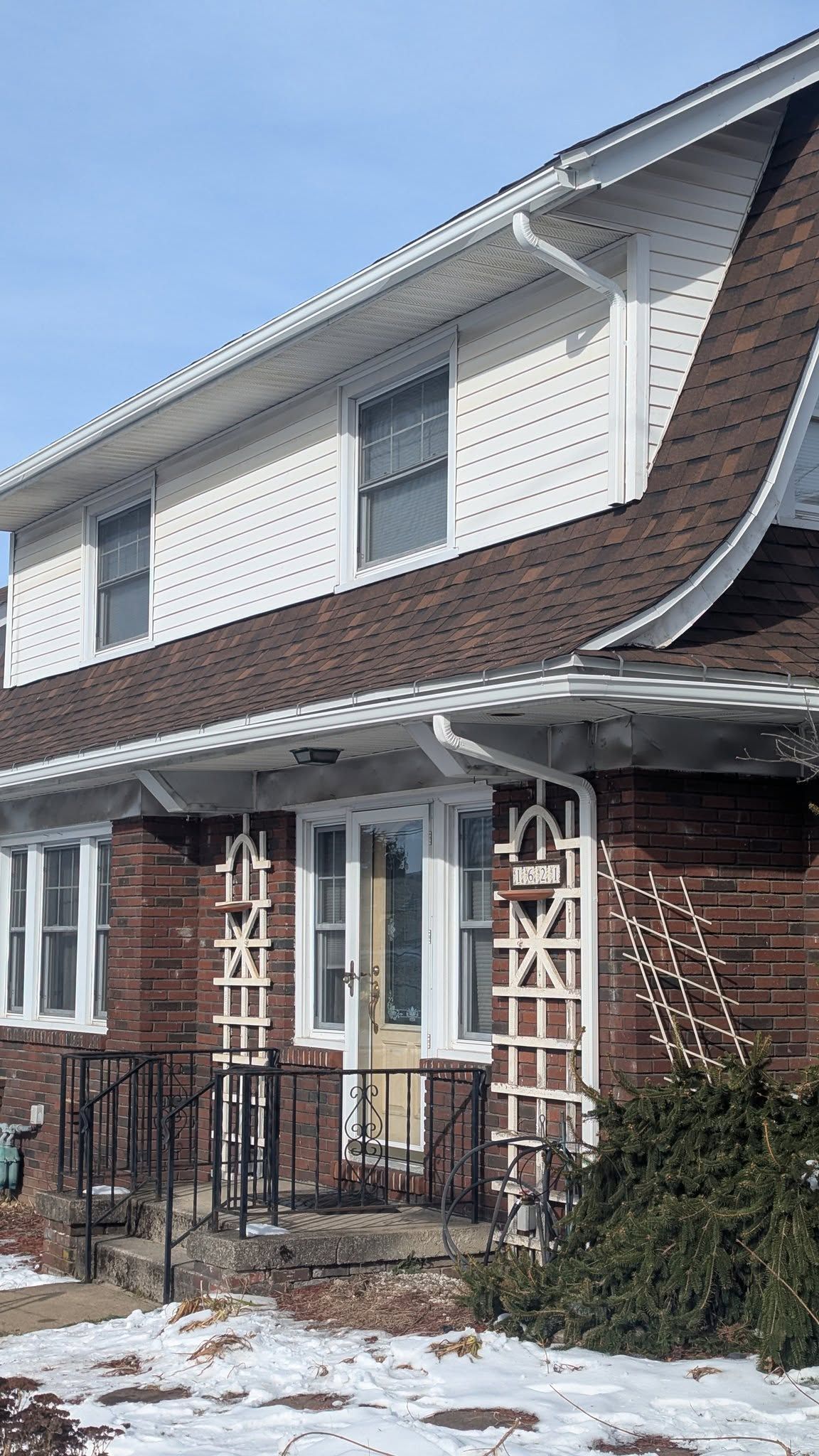 Snow-dusted brick house with white siding, front porch, and decorative trellis supports