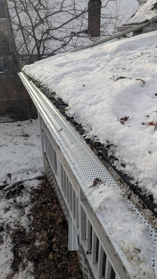 Snow-covered roof edge with white gutter and bare trees in the background