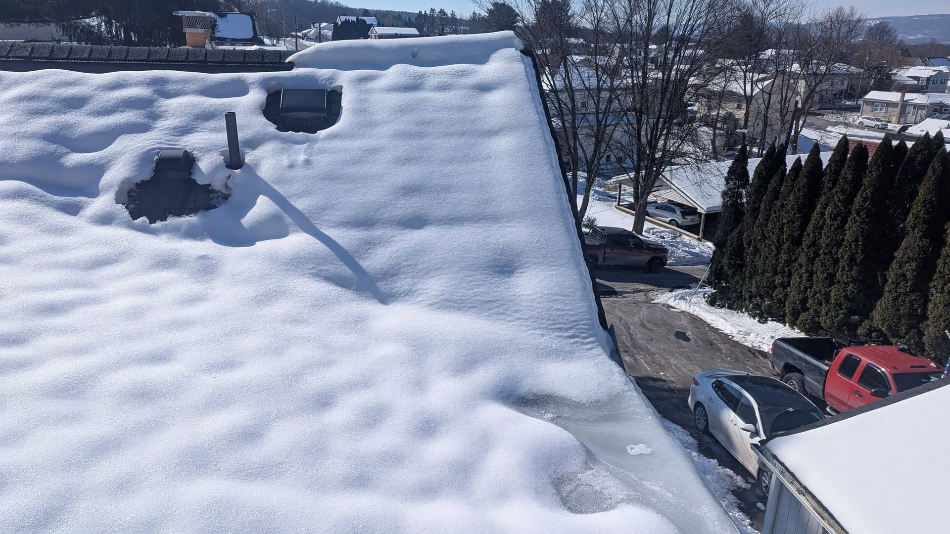Snow-covered rooftop with two vents overlooking a residential street and parked cars on a sunny winter day