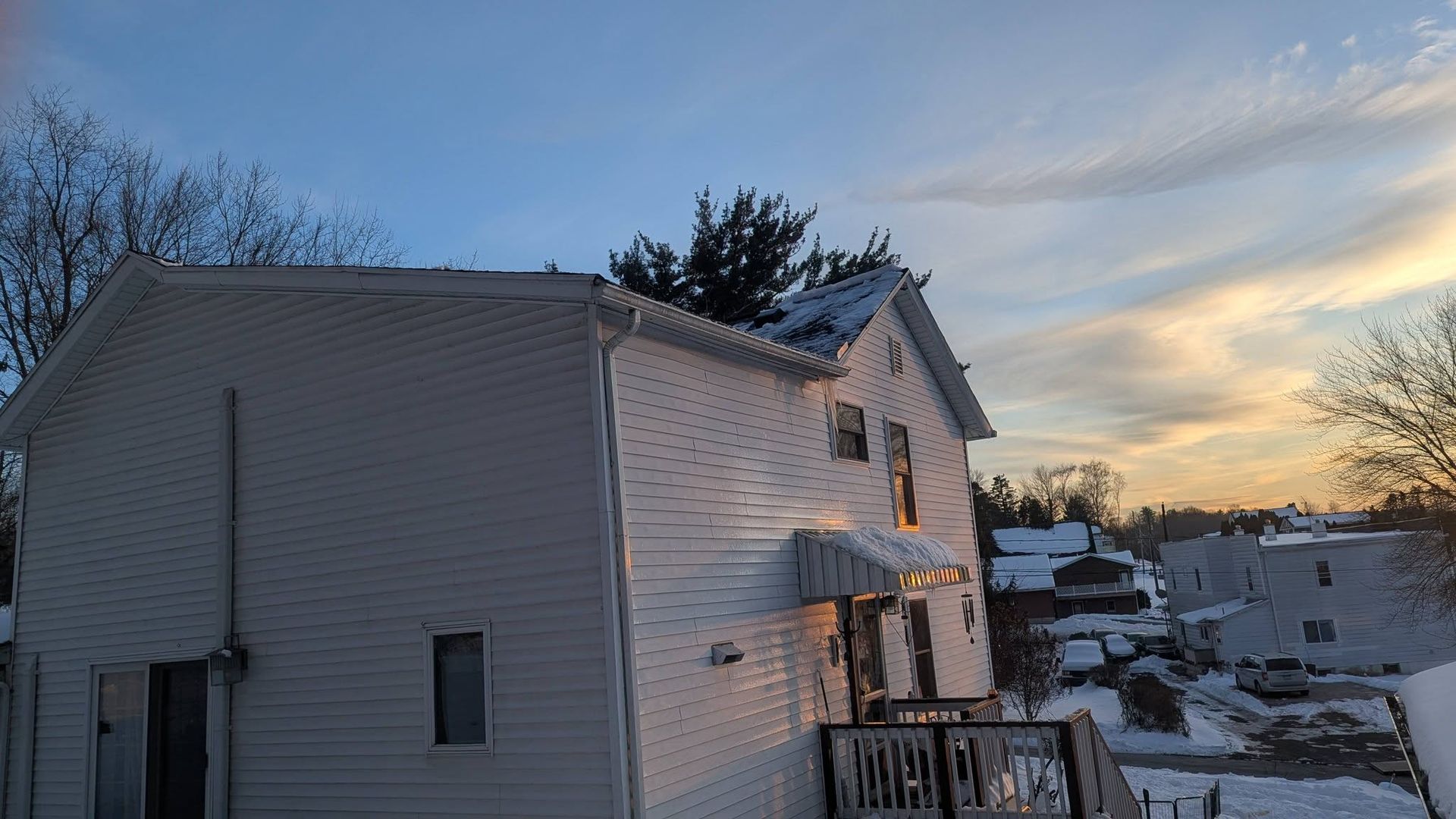 Snow-covered house at sunset with damaged siding and a winter residential neighborhood in the background