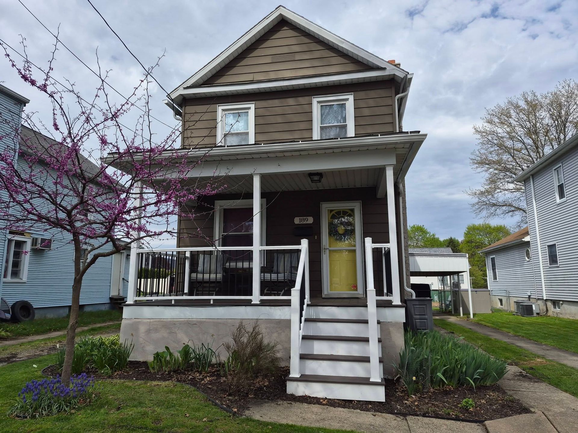 Small two-story house with a front porch, white railing, and yellow door on a cloudy day.