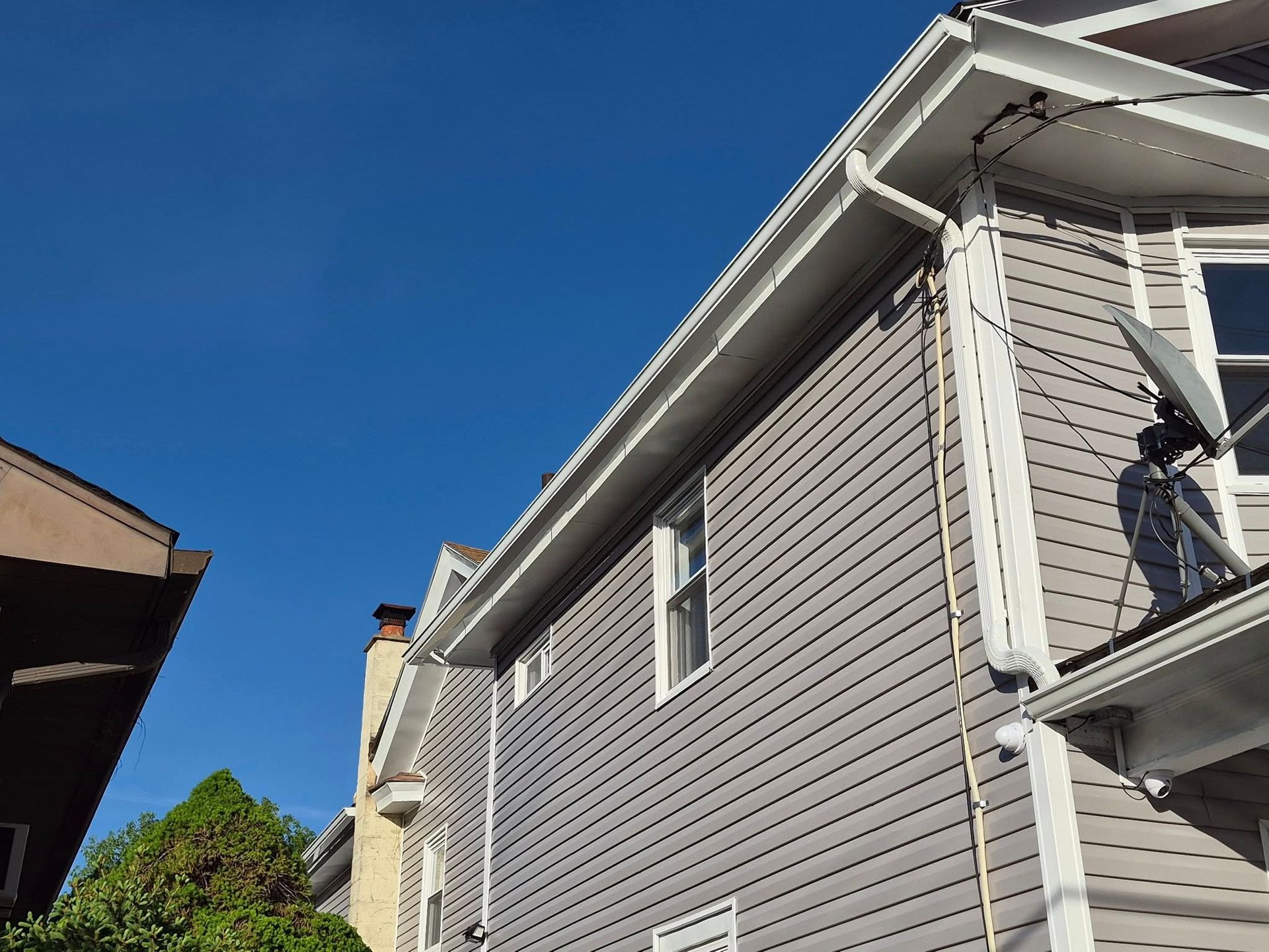 Gray house siding and roof eaves under a bright blue sky, with neighboring buildings at the edges