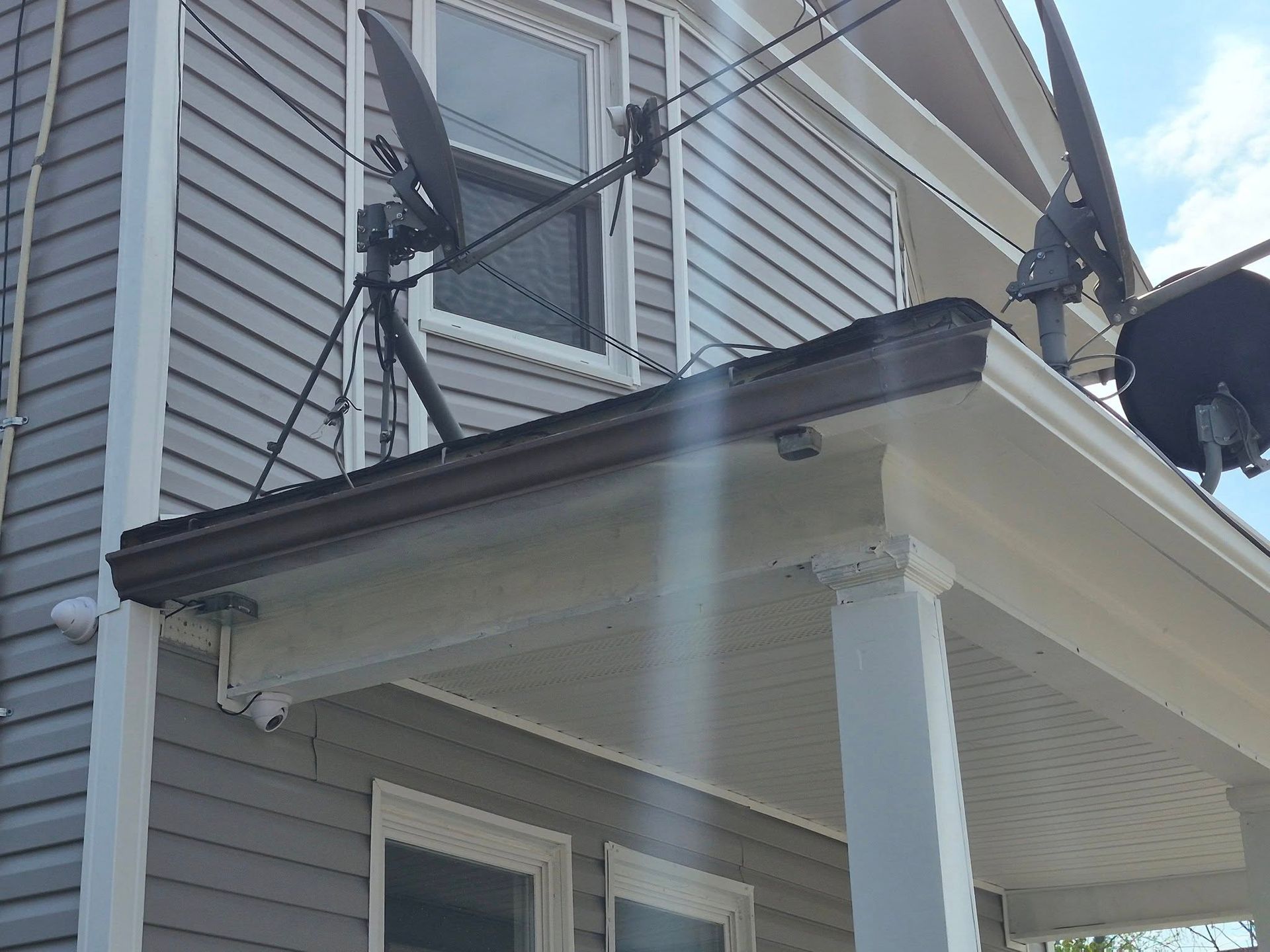 Two satellite dishes mounted on a house roof above a white porch with gray siding.