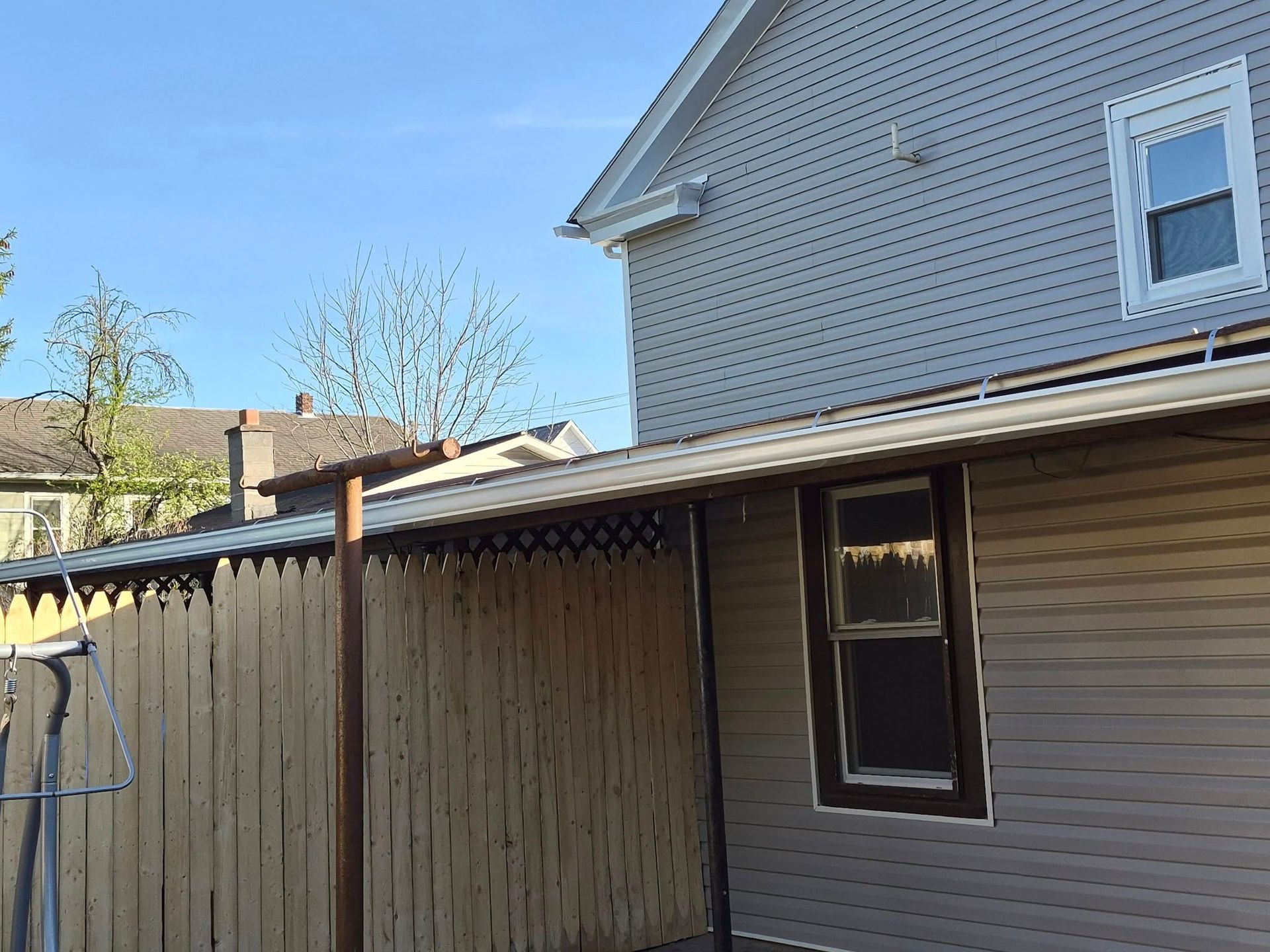 Backyard side of a house with beige siding, wooden fence, and covered patio roof under blue sky