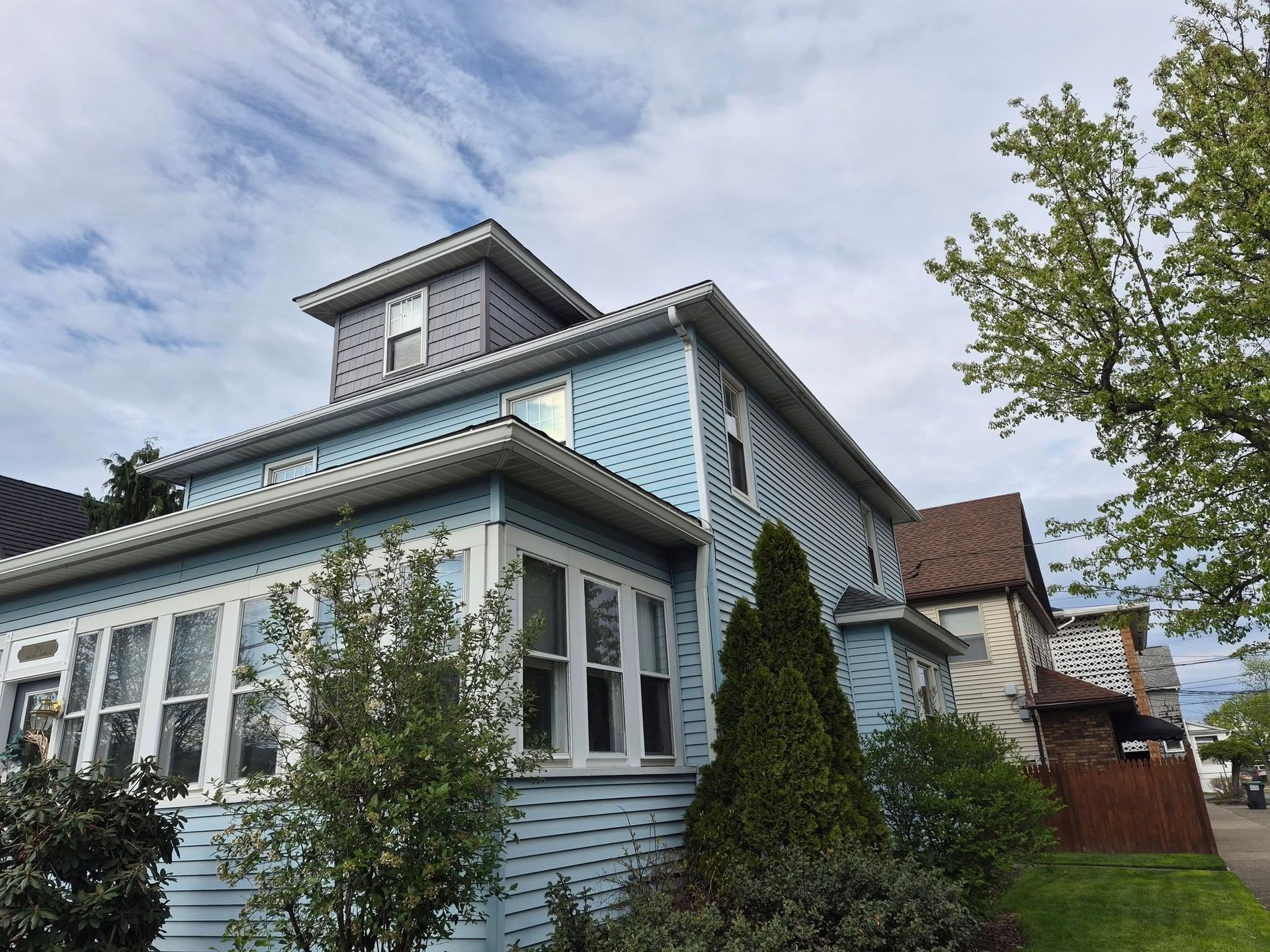 Light blue house with white trim, dormer windows, and a front garden under a cloudy sky