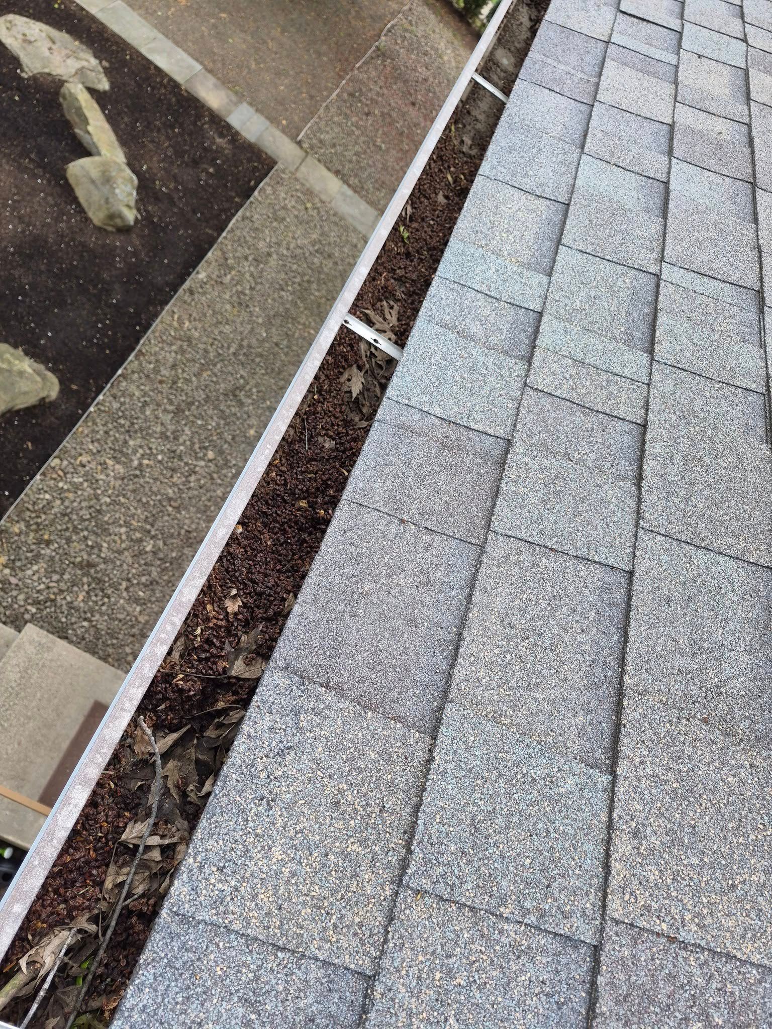 Sloped gray shingle roof beside a gutter and a tan stone walkway with fallen leaves and debris.