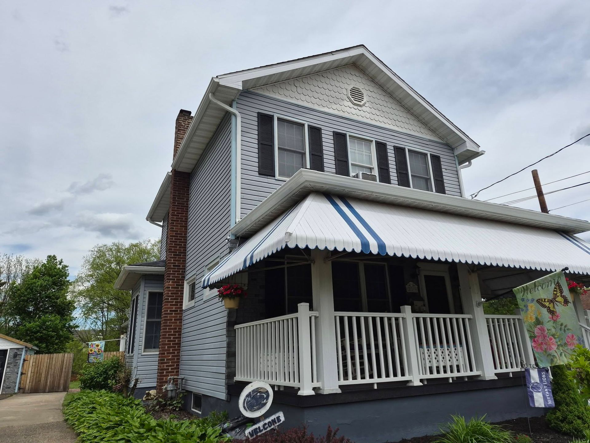 Two-story white house with a front porch, striped awning, and brick chimney on a cloudy day