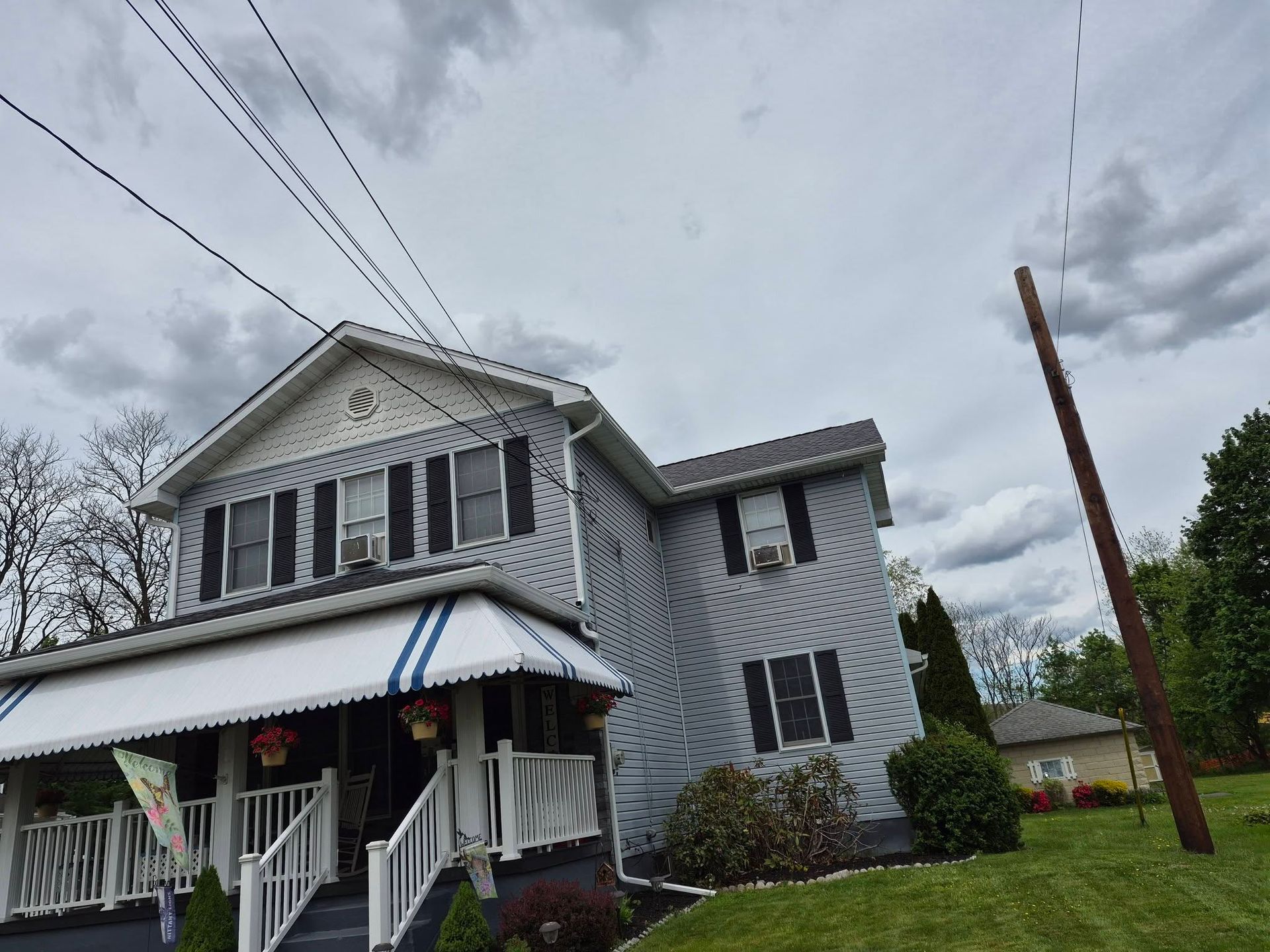 Gray two-story house with black shutters, striped awning, and porch on a cloudy day