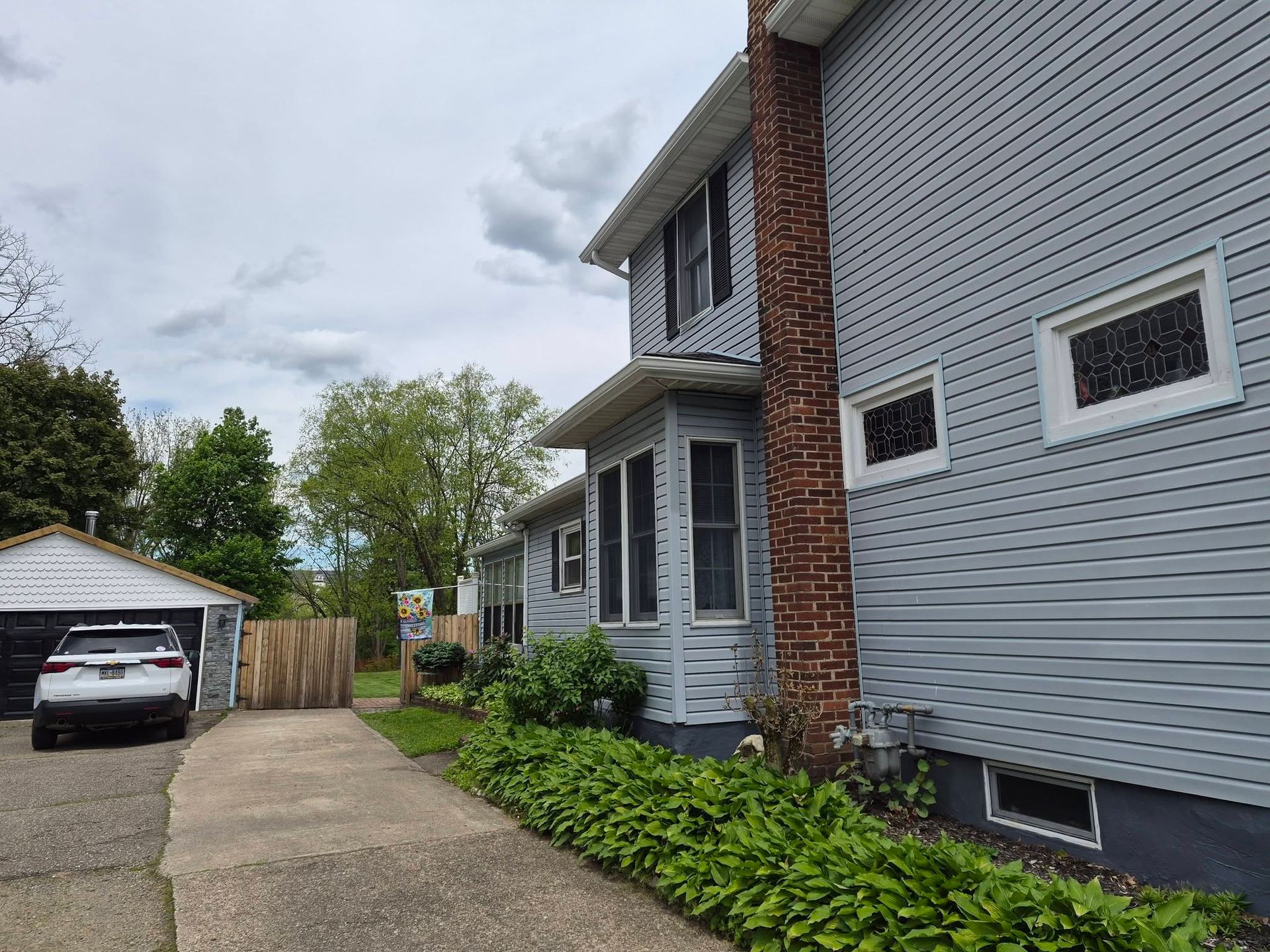 Gray two-story house with brick chimney, driveway, detached garage, and green shrubs along the side.